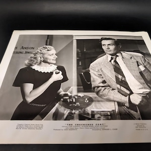 May include: Black and white promotional image from the film "The Checkered Coat." A woman in a dark dress with a white collar examines jewelry. A man in a plaid jacket and tie stands nearby. The image includes text from the film.