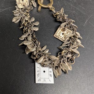 May include: A silver-toned bracelet with leaf charms and watch face accents. The bracelet features a toggle clasp and is composed of multiple chains. The watch faces have Roman numerals. The bracelet is on a black surface.