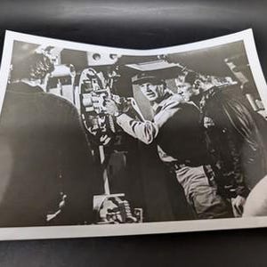 May include: Black and white photograph of a group of men operating machinery. One man in a uniform and hat is focused on the equipment, while others observe. The scene appears to be indoors, possibly a military setting.