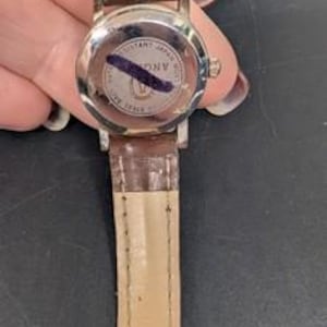 May include: A silver-toned wristwatch with a brown leather band. The watch face is not visible. The back of the watch has a circular design with text and a dark blue mark. The band is a medium brown color.