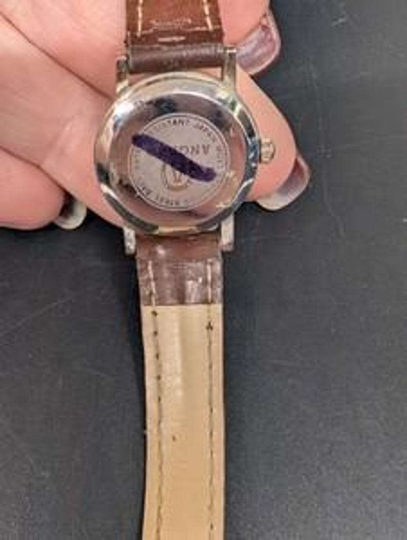 May include: A silver-toned wristwatch with a brown leather band. The watch face is not visible. The back of the watch has a circular design with text and a dark blue mark. The band is a medium brown color.