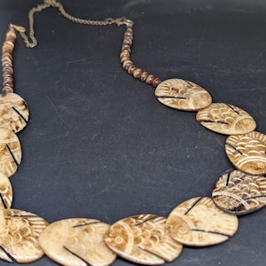 May include: A necklace with oval, beige pendants, each etched with a fish design. The pendants are linked by a brown beaded strand and a brass-coloured chain. The necklace is set against a dark background.