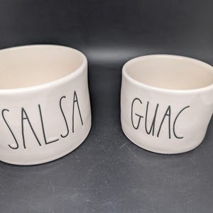 Pode incluir: Duas tigelas de cerâmica branco-sujo. A tigela maior tem a palavra "SALSA" em letras pretas. A tigela menor tem a palavra "GUAC" em letras pretas. Ambas as tigelas têm um design simples e moderno, provavelmente para servir molhos.