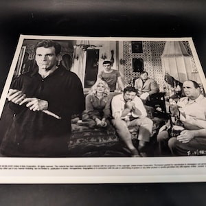 May include: Monochrome photograph of a group of people in a room. A man in a dark top holds a long, thin object. Several other people are seated or standing in the background. The image is a vintage movie still.