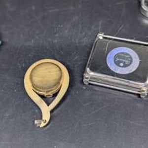 May include: A gold-colored, oval-shaped compact mirror with a decorative handle. Next to it is a small, rectangular, clear case with a blue and white circular design. The items are on a dark surface.