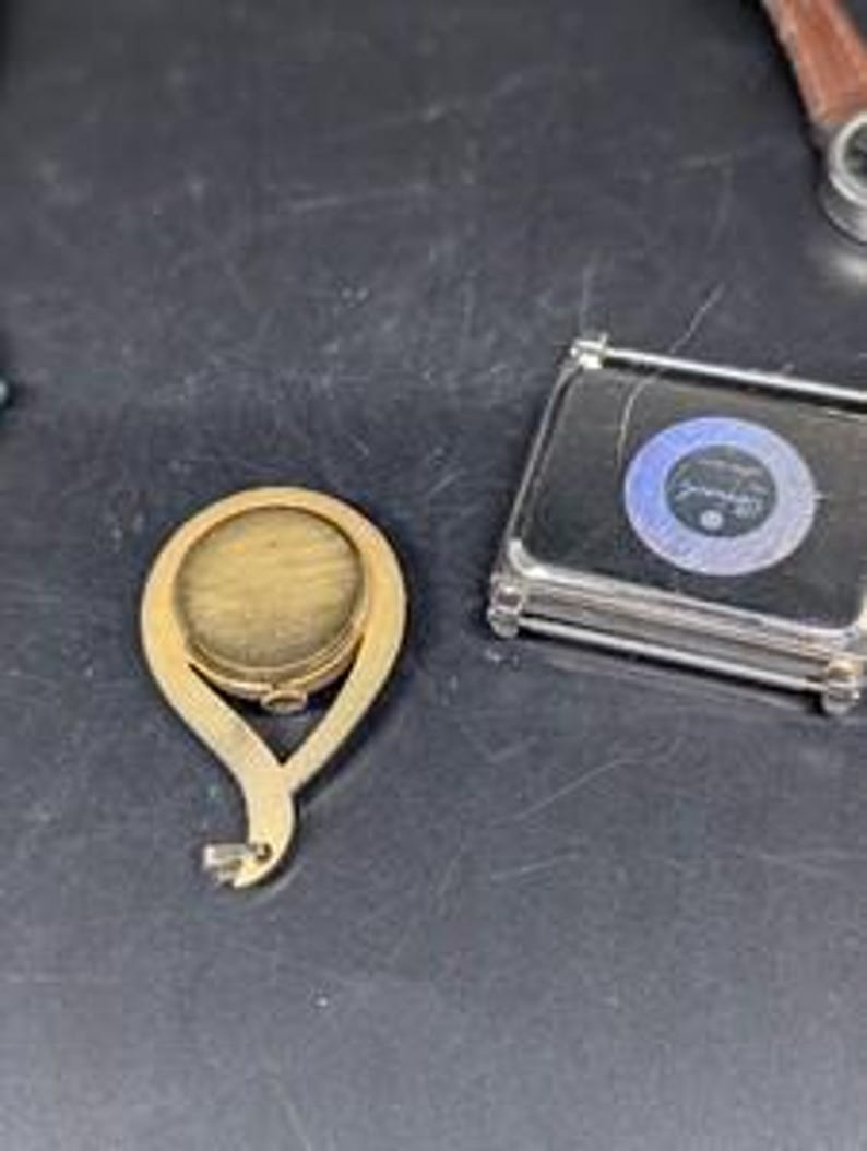 May include: A gold-colored, oval-shaped compact mirror with a decorative handle. Next to it is a small, rectangular, clear case with a blue and white circular design. The items are on a dark surface.
