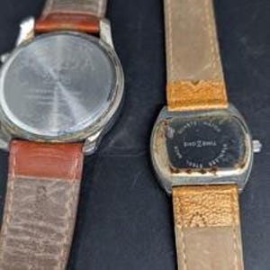 May include: Two wristwatches with leather bands are displayed. One watch has a silver case and a brown leather band. The other watch has a rectangular face and a light brown leather band. Both watches show signs of wear.