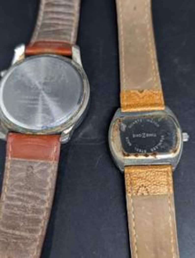 May include: Two wristwatches with leather bands are displayed. One watch has a silver case and a brown leather band. The other watch has a rectangular face and a light brown leather band. Both watches show signs of wear.