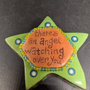 May include: A green star-shaped decorative item with the text "there's an angel watching over you." The star has blue and white polka dots and a beaded border in yellow and green. The center is orange.