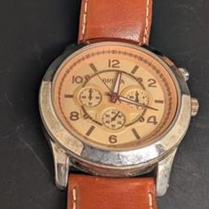 May include: A close-up of a wristwatch with a brown leather band and a silver-colored case. The watch face is tan with black numerals and three smaller dials. The watch hands are black. The watch is on a black surface.