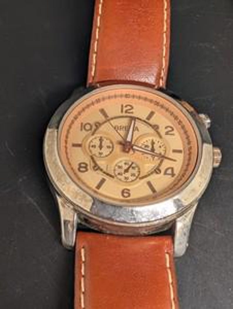 May include: A close-up of a wristwatch with a brown leather band and a silver-colored case. The watch face is tan with black numerals and three smaller dials. The watch hands are black. The watch is on a black surface.