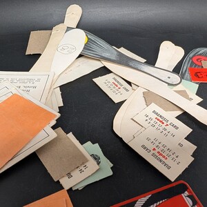 May include: A collection of vintage ephemera, including paper cutouts, cards, and printed ephemera. The items include a silver-colored knife, cards with the text "DIAGNOSIS CARD", and other paper pieces with text and numbers. The items are scattered on a dark surface.