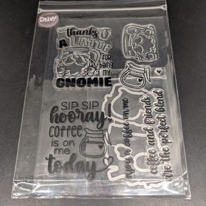 May include: Clear stamp set featuring coffee-themed designs and phrases. Includes a gnome with a coffee cup, coffee pots, and text that reads "thanks a latte," "sip sip hooray," and "enjoy a coffee on me."