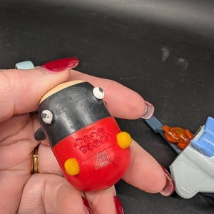May include: A small, rounded toy figure with a black body, red lower half, and yellow accents. The toy has white eyes and a light tan head. The bottom of the toy is red and has the text "JAKKS CHINA 0128BT".