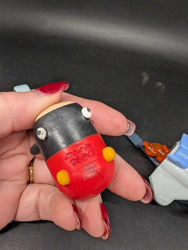 May include: A small, rounded toy figure with a black body, red lower half, and yellow accents. The toy has white eyes and a light tan head. The bottom of the toy is red and has the text "JAKKS CHINA 0128BT".