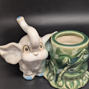 May include: A ceramic elephant figurine in white and light gray with blue accents. The elephant is next to a green planter with a textured design. The planter has a round opening. The figurine is on a black surface.
