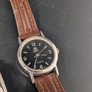 May include: A silver-toned analog watch with a black face and brown leather band. The watch face displays the brand name "ANGARO" and the word "QUARTZ". The watch band is stitched with a lighter brown thread.