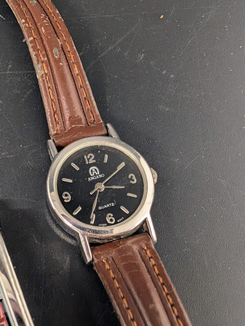 May include: A silver-toned analog watch with a black face and brown leather band. The watch face displays the brand name "ANGARO" and the word "QUARTZ". The watch band is stitched with a lighter brown thread.