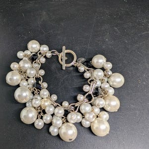 May include: A silver-toned bracelet with a toggle clasp, adorned with clusters of white and off-white faux pearls of varying sizes. Small black beads are interspersed among the pearls, adding contrast. The bracelet is on a dark surface.