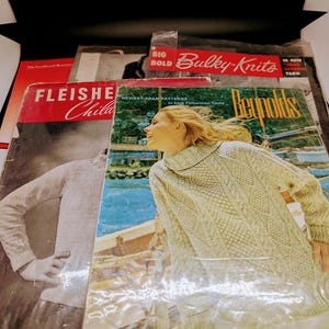 May include: A selection of vintage knitting pattern books. The covers display images of sweaters and the text "Bulky-Knits", "Fleishe Child", and "Reynolds". The books are in various colors, including red, green, and white.