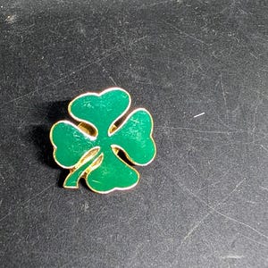 May include: A green enamel shamrock pin with a gold-colored outline. The four-leaf clover design is a symbol of good luck and is often associated with St. Patrick's Day.