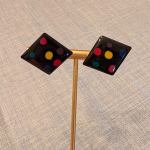 May include: A pair of black, diamond-shaped earrings with colorful polka dots in red, yellow, purple, and teal. The earrings are displayed on a gold-colored stand, showcasing their glossy finish and playful design. These earrings are a fun accessory.
