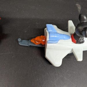 May include: A toy jet plane with a blue cockpit and orange exhaust. The jet has a gray body and a black figure with red shoes is seated in the cockpit. The jet is on a black surface.