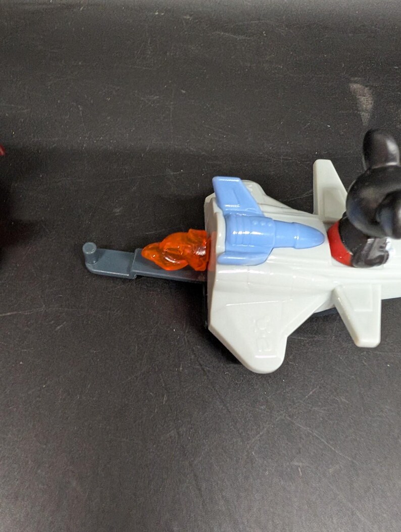 May include: A toy jet plane with a blue cockpit and orange exhaust. The jet has a gray body and a black figure with red shoes is seated in the cockpit. The jet is on a black surface.