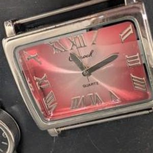 May include: A rectangular silver-toned quartz watch with a red face and Roman numeral hour markers. The watch has a silver band and the word "Shock" is printed on the face.