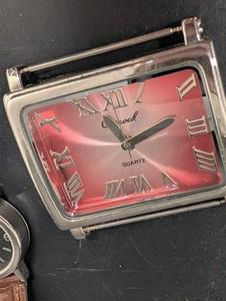 May include: A rectangular silver-toned quartz watch with a red face and Roman numeral hour markers. The watch has a silver band and the word "Shock" is printed on the face.