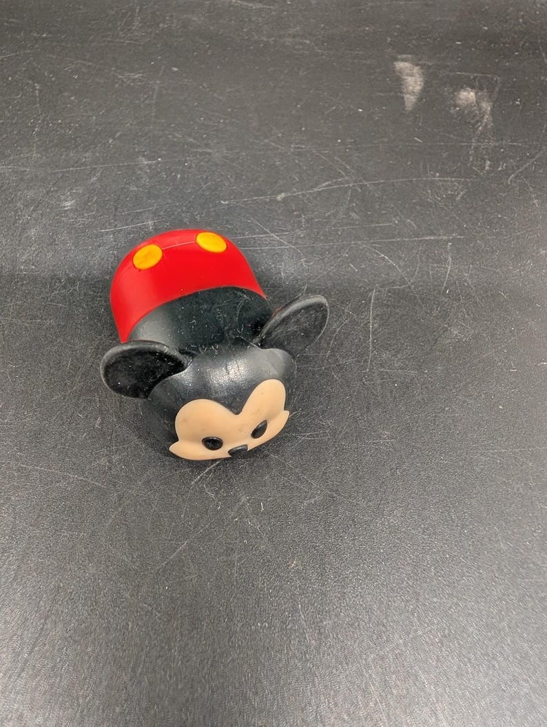 May include: A small, black and red Mickey Mouse figurine with yellow accents. The figurine has large black ears, a red head with two yellow circles, and a tan face. The toy is lying on a dark surface.