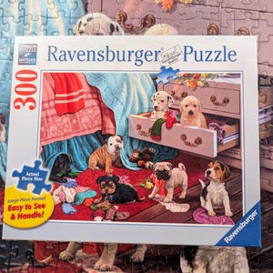 May include: A Ravensburger 300-piece jigsaw puzzle box featuring a colorful illustration of various puppies playing in a room with a wooden dresser, a red rug, and blue fabric. The box includes text indicating the piece count and puzzle brand.