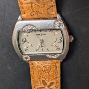 May include: A silver-toned wristwatch with a light-colored face and a brown leather band. The watch face displays the text "TIME ZONE QUARTZ" and features the numbers 3 and 9. The band is decorated with a floral pattern.