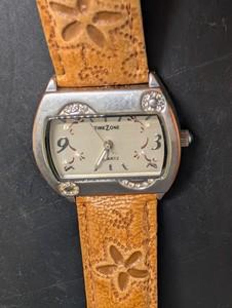May include: A silver-toned wristwatch with a light-colored face and a brown leather band. The watch face displays the text "TIME ZONE QUARTZ" and features the numbers 3 and 9. The band is decorated with a floral pattern.