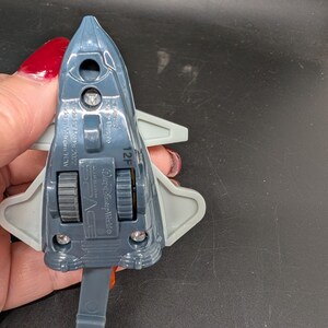 May include: A gray and blue toy space shuttle with gray wings and a pull-down handle. The toy has two black wheels and the words "Space" and "Disney World" printed on it.