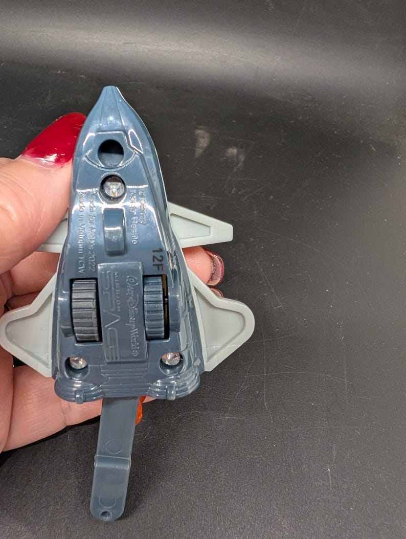 May include: A gray and blue toy space shuttle with gray wings and a pull-down handle. The toy has two black wheels and the words "Space" and "Disney World" printed on it.