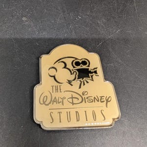 May include: A gold-colored pin featuring the Walt Disney Studios logo. The pin has a rectangular shape with rounded edges. The top of the pin features a silhouette of Mickey Mouse and a film camera. The text "The Walt Disney Studios" is inscribed.