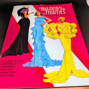 May include: A vintage book cover with a pink background and illustrations of three women in elegant gowns. The text reads "Great Fashion Designs of the Thirties" and lists designers like Schiaparelli and Molyneux.