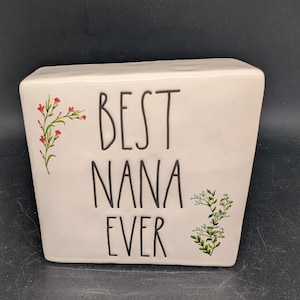 May include: A white ceramic rectangular vase with the words "BEST NANA EVER" in black lettering. The vase is decorated with floral accents in red and green.