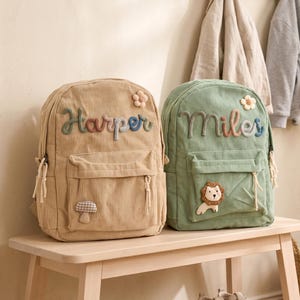 May include: Two corduroy backpacks, one tan and one green, sit on a wooden bench. The tan backpack has the name "Harper" embroidered in colorful thread, and the green backpack has "Miles". Both have a front pocket and decorative patches.