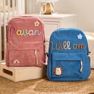 May include: A pink corduroy backpack with the name Pavan in colorful letters, a flower patch, and a rainbow patch. Beside it, a blue corduroy backpack with the name William in colorful letters, a flower patch, and a hedgehog patch.