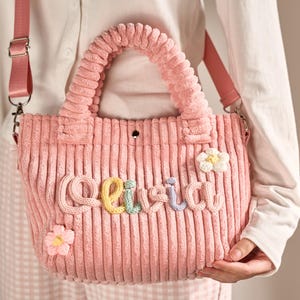 May include: A pink corduroy tote bag with a top handle and a detachable shoulder strap. The bag features the name "Olivia" in colorful, raised letters and is adorned with fabric flower accents. The bag is a soft pink color.