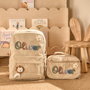May include: A beige corduroy backpack and matching lunch bag, both embroidered with the name "Oliver" in colorful letters. The backpack features a lion applique and a flower. A wooden chair with bunny ear backrest and a wooden bookshelf are also in the image.