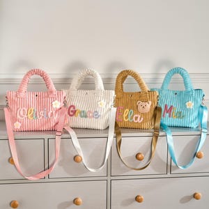 May include: Four corduroy tote bags in pink, white, tan, and light blue, each featuring a matching adjustable shoulder strap, a top handle, and a personalized name embroidered on the front. The bags are decorated with small fabric flower or bear appliques.