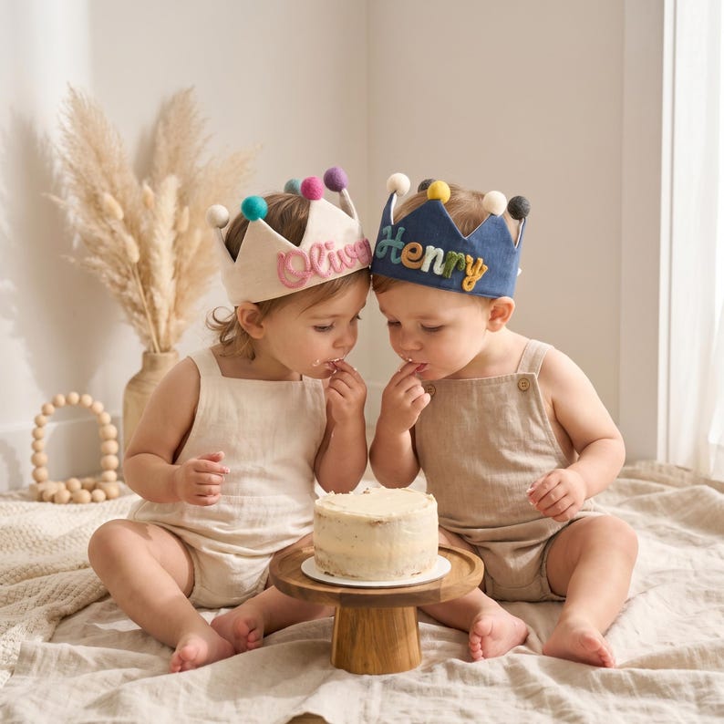 May include: Two children wearing felt crowns with colorful pom-poms and names embroidered on them. One crown is cream-colored with the name "Olivia" in pink, and the other is navy blue with "Henry" in colorful letters. They are eating cake.