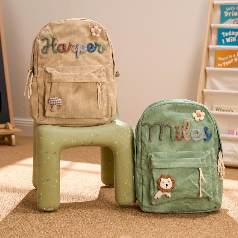 May include: Two small, personalized backpacks. The beige backpack has the name "Harper" embroidered on the front with a mushroom and flower applique. The green backpack has the name "Miles" embroidered on the front with a lion and flower applique.