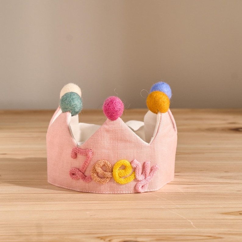 May include: A pink fabric crown with the name "Zoey" embroidered in colorful thread. The crown is decorated with colorful pom-poms and sits on a wooden surface. The crown is designed for a child.