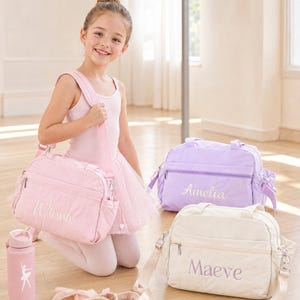 May include: Three rectangular duffel bags in light pink, lavender, and cream, each featuring a personalized name embroidered on the front. The bags have adjustable shoulder straps, side pockets, and zippered closures. A pink water bottle and ballet slippers.
