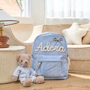 May include: A light blue corduroy backpack with a white rope name tag that says "Adora" and a small gray bow. The backpack is sitting on a light brown surface next to a teddy bear wearing a blue and white striped shirt.