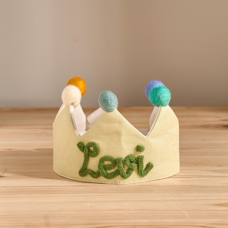 May include: A light yellow fabric crown with the name "Levi" embroidered in green. The crown is decorated with colorful pom-poms in yellow, white, teal, and blue. The crown is designed for a child.